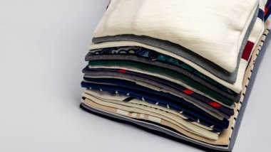 Katlanmış kıyafet yığınını gösteren, çeşitli doku, renk ve desenlere sahip yakın plan bir fotoğraf. Yığında gömlekler ve kazaklar yer alıyor ve görsel açıdan çekici bir görüntü yaratıyor. Görüntü iyi aydınlatılmış, farklı kumaşları vurguluyor ve 