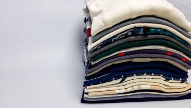 Düzgünce katlanmış, çeşitli renkler, dokular ve desenler içeren uzun bir giysi yığınının yakın çekimi. Giysiler organize bir yığın halinde düzenlenmiş. Yumuşak ışıklandırma ve nötr arkaplan temiz ve minimalist 