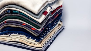 Düzgünce istiflenmiş katlanmış giysi parçalarının yakın plan çekimi. Kıyafetler farklı renk ve desenlerde, görsel olarak çekici bir kompozisyon oluşturuyor. Tarafsız bir arkaplan ile iyi ışıklandırılmış görüntü, dokuyu ve ayrıntıları vurguluyor