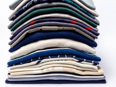 Yakın plan çekimde düzgünce dizilmiş bir yığın kıyafet sergilenir. Koleksiyonda çeşitli renkler, desenler ve süveterler, gömlekler ve diğer giyim ürünleri yer alıyor. Temiz beyaz arkaplan çeşitliliği vurgular.