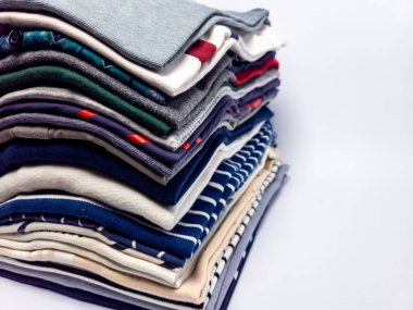 Temiz beyaz bir arkaplana karşı düzgünce dizilmiş katlanmış kıyafetlerin yakın plan çekimi. Giysiler farklı renk ve desenlerde, tişört, gömlek ve süveter gibi bir takım elbiseler olduğunu gösteriyor. Resim temizliği ve organizasyonu vurguluyor