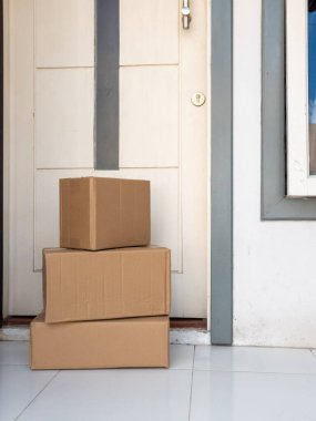 Üç karton kutu, gümüş saplı ve anahtar delikli beyaz bir kapının önündeki fayanslı bir verandaya istiflenir. Kutular düz kahverengi, yeni bir doğum olduğunu gösteriyor. Görüntü, kapı ve kutuların yakın çekimini gösteriyor. Sahne online alışveriş öneriyor.