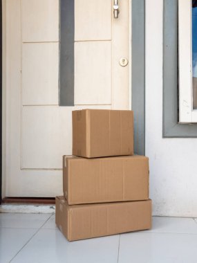 Üç kahverengi karton kutu yığını bir evin fayanslı verandasında duruyor, tam beyaz ön kapının önünde. Paketler ev sahibini beklerken teslimat tamamlanmış gibi görünüyor. Sahne online alışveriş, ev teslimatı veya