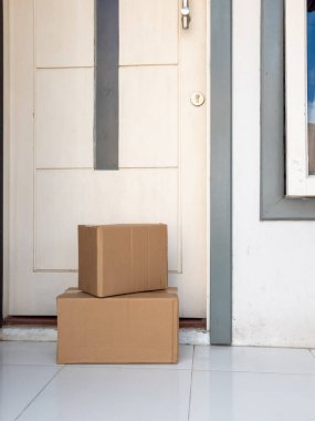 İki karton kutu beyaz bir kapının önündeki fayanslı verandada duruyor. Kutular alıcının onları açması için hazır. Kapının dikey koyu gri aksanı olan modern bir tasarımı var. Sahne online alışveriş yapmayı öneriyor..