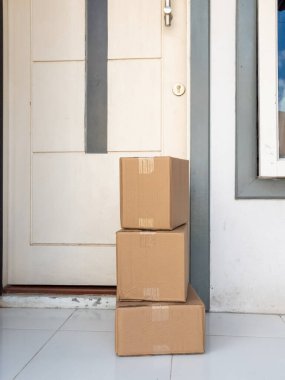 Üç tane istiflenmiş karton kutu bir evin ön verandasında duruyor, alınmayı bekliyorlar. Kutular beyaz bir kapının önüne düzgünce yerleştirilmiş. Bu da yeni bir teslimat olduğunu gösteriyor. Görüntü çevrimiçi alışveriş, e-ticaret ve ev teslimatı hizmetleri kavramlarını iletir