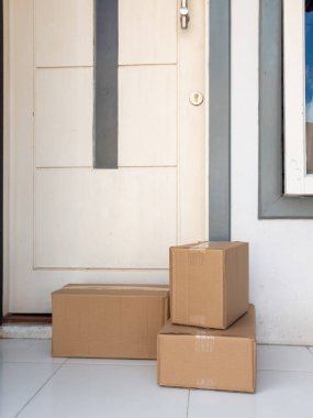 Üç kahverengi karton kutu, dikey koyu gri aksanlı beyaz bir ön kapının önündeki fayanslı bir kapı eşiğine istiflenir. Sahne, çevrimiçi alışveriş ve ev dağıtım hizmetlerinin kolaylığını vurgulayan bir teslimat veya paket ulaşım öneriyor.