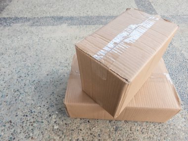 Yüksek açılı iki karton kutunun desenli beton bir yüzeyde görüntüsü. Kutular bantla mühürlenmiş ve nakliye ya da teslimat için hazır görünüyor. Görüntü nötr tonlar ve çeşitli parçalar için uygun basit bir kompozisyon içerir. 
