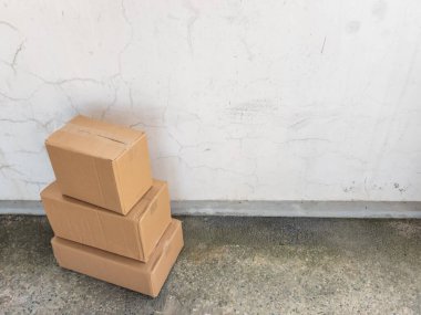 Üç kahverengi karton kutu, yıpranmış beyaz bir duvara karşı üst üste istiflenir. Kutular hafifçe açılı, dinamik bir bileşim oluşturuyor ve beton zemin doku ekliyor. Görüntü iyi aydınlatılmış, doğal ışık vurgulanıyor.