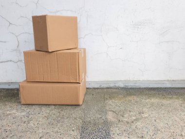 Kırık beyaz bir duvara karşı üst üste istiflenmiş üç kahverengi karton kutu. Kutular desenli beton bir yüzeye yerleştirilmiş. Görüntü basit bir sahneyi vurgular, muhtemelen nakliye, taşınma veya depolama ile ilgilidir. Yıpranmış duvar ve