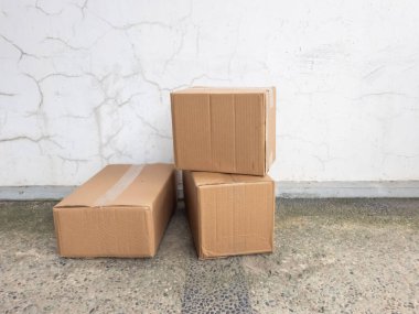 Üç karton kutu istiflenmiş ve belirgin çatlakları olan desenli beyaz bir duvara karşı yerleştirilmiş. Kutular küçük çakıl taşlarıyla beton bir yüzeyde duruyor. Işıklandırma bile nötr ve faydacı bir estetik yaratıyor. İllüstratin için mükemmel.