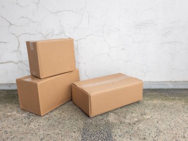 Değişen boyutlarda üç mukavva kutu benekli beton bir yüzey üzerinde çatlak desenli beyaz bir duvara karşı duruyor. Kutular kapalı ve bantlı, nakliye ya da depo için hazır olduklarını gösteriyor. Sahne basit ve tarafsız, tema içeriyor.