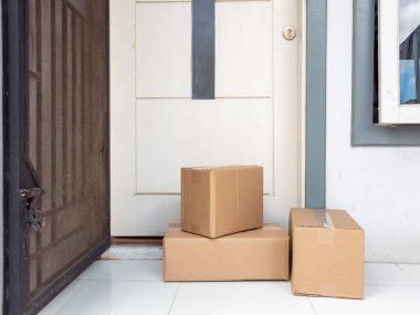 Birkaç karton kutu kapının önünde duruyor, bu da yeni bir teslimatı gösteriyor. Kutular düzgünce bir evin girişine yerleştirilmiş, bu da online alışveriş ya da paket teslimatı olduğunu gösteriyor. Açık güvenlik kapısı içeriyi bir anlığına gösteriyor. Bu resim c