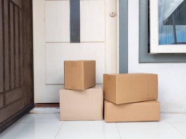 Birkaç karton kutu ön kapının yanına istiflenmiş. Bu da yeni bir teslimat olduğunu gösteriyor. Kutular farklı boyutlarda, içerideki çeşitli nesneleri gösteriyor. Sahne aydınlık, temiz beyaz fayansları ve kutuların nötr tonlarını vurguluyor. 