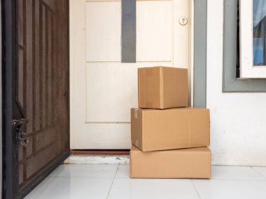 Kapalı bir kapının önündeki beyaz bir verandaya istiflenmiş üç kahverengi karton kutu, muhtemelen teslimattan sonra. Görüntü, online alışveriş ve ev teslimatlarının ortak bir sahnesini yakalıyor. Sahne doğal ışıkla aydınlatıldı. Ön planda, 