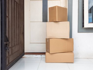 Dört karton kutu bir evin kapısında fayanslı bir zeminde duruyor. Kutular çeşitli boyutlarda, ve kapı biraz aralık. Olay yeri, yeni teslimat ya da online alışveriş olduğunu gösteriyor. Kompozisyon, ev teslimatının uygulanabilirliğini vurguluyor