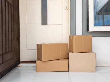 Beyaz bir kapının önündeki verandada duran karton kutular. Kutular farklı boyutlarda. Birden fazla teslimat olduğunu gösteriyor. Görüntü, çevrimiçi alışveriş ve ev dağıtım hizmetlerinin kolaylığını vurguluyor. Sahne ışıl ışıl parlıyor.