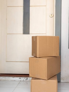 Beyaz bir kapının önündeki kapı eşiğine istiflenmiş üç karton kutu bekleyen bir teslimatı gösteriyor. Paketlere ve sevkiyat beklentilerine odaklan. Basit kompozisyon, doğal ışıklandırma. Olay yeri aydınlık, kutular temiz.,