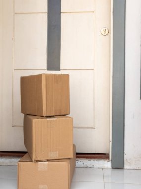 Üç karton kutudan oluşan bir deste verandada oturmuş, alınmayı bekliyor. Kutular çeşitli boyutlarda ve koli bandı var. Gri süslemeli ve anahtar delikli beyaz bir kapının önüne yerleştirilmişler. Fotoğraf iyi aydınlatılmış ve şarküteriye odaklanmış.