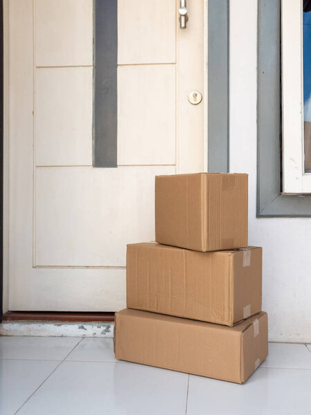 Three cardboard boxes stacked on top of each other are sitting on a tiled front porch. The boxes are brown and have tape on them. Behind the boxes is a white door with a silver handle and a keyhole. The scene suggests a delivery or online shopping.