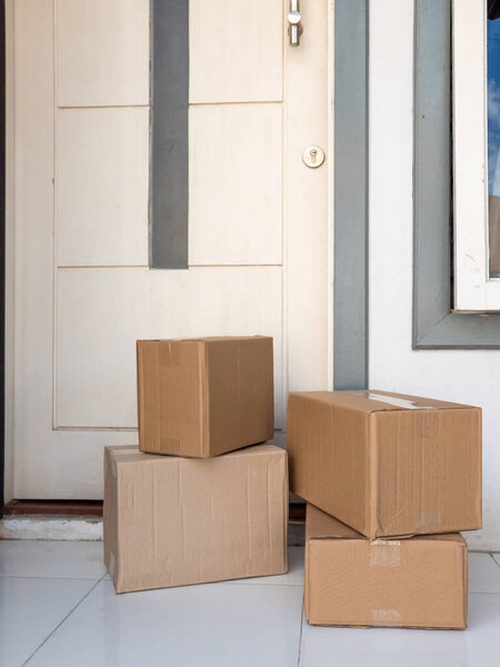 A pile of brown cardboard boxes are stacked on a tiled porch in front of a white door. The boxes appear to have been delivered. The scene is bright and natural light illuminates the scene. The composition is simple, focusing on the boxes and the door