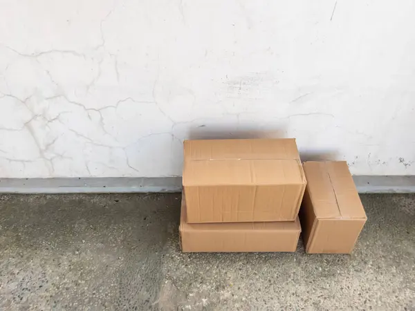 Üç karton kutu desenli, çatlamış beyaz bir duvara karşı istiflenmiş. Kutular çeşitli boyutlarda ve yeni gibi görünüyor. Zemin benekli beton bir yüzey. Işıklandırma doğal, ve genel sahne bir bekleme ya da delive hissi taşır