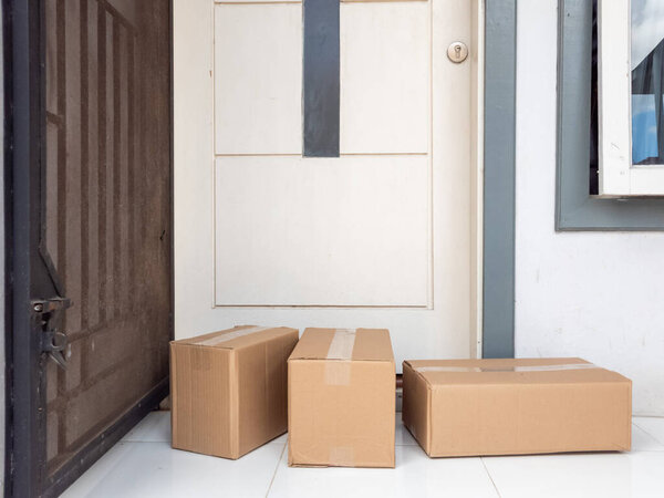Three brown cardboard boxes sit on a tiled porch in front of a white door, indicating a recent delivery. The image captures the convenience of online shopping and package delivery services. The scene is brightly lit, suggesting a sunny day, and the c