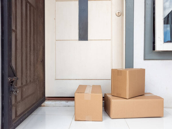 Three brown cardboard boxes sit on a tiled porch outside a cream-colored front door. The image shows a delivery of packages left on a doorstep, with an open security gate and window visible. The boxes are sealed and ready for the homeowner to retriev
