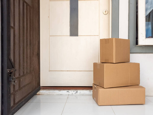 Three brown cardboard boxes stacked on a tiled porch, near a white door. The door is partially open, and a security screen door is visible. The scene depicts a delivery of packages at a home, likely representing online shopping or a mail delivery. Th