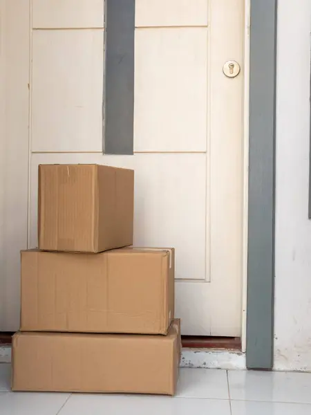 Üç kahverengi karton kutu beyaz bir ön kapının önüne istiflenmiş. Kutular farklı boyutlarda ve yeni teslim edilmiş gibi görünüyor. Arka planda biraz yıpranmış bir kapı ve anahtar deliği var. Zemin kiremit kaplı ve sahne bir fahişeyi işaret ediyor.