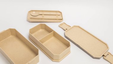 Beyaz bir arka planda bej bir bento kutusu parçalanmış olarak gösterilir. İçinde bir kapak, birkaç kompartıman ve kaşık ve yemek çubukları olan bir tepsi var. Resim yemek kabının tasarımı, dokusu ve özelliklerini vurguluyor.