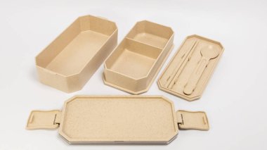 Beyaz arka planda bej bento seti sunuluyor. Set, kompartımanlı bir dikdörtgen kutu, mandallı bir kapak ve içinde yemek çubukları ve bir kaşık bulunan daha küçük bir kap içeriyor. Ögeler tasarımlarını ve işlevlerini göstermek için düzenlenmiştir
