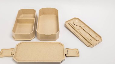 Kapaklı bir bej bento kutusu ve iki ayrı bölme, yanında da çatal bıçak takımı. Beslenme çantası açık, içini gösteriyor. Set yemek çubukları ve bir kaşık içeriyor. Eşyalar beyaz bir yüzeye yerleştiriliyor. Yemek fotoğrafçılığı için ideal.