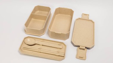 Açık bej bir bento seti, kaşık ve yemek çubuklarıyla, paketlenmiş öğle yemeği için hazır. Sette iki yemek kabı, bir kapak ve çatal bıçak için bir tepsi yer alıyor. Ögeler beyaz bir yüzeye dizilir, materyalin doğal dokusunu vurgular.. 
