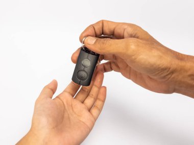 Yakın plan çekimde bir çift el temiz beyaz bir arka planla araba anahtarlığını değiş tokuş ederken görülüyor. Görüntü, bir arabanın mülkiyet, satın alma veya kiralanmasını sembolize ediyor. Odak keskin, iyi ışıklandırmayla anahtarlığı ve elleri vurguluyor.