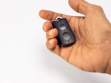 Yakından çekilen bir fotoğraf, parlak, siyah bir anahtarlık tutan bir el sergiler. Anahtarlıkta iki düğme ve muhtemelen bir araba için bir güç sembolü var. El iyi ışıklandırılmış, görünür deri dokusuyla, temiz beyaz bir zemin üzerine kurulmuş, anahtarı ve onun 