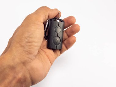 Siyah bir arabanın anahtarlığını beyaz arka planda tutan bir elin yakın çekimi. Anahtarlığın iki tuşu var ve metal bir yüzük görülebilir. El iyi aydınlatılmış, deri dokusu ve ayrıntıları sergiliyor. Kompozisyon basit. Doğuma odaklanıyor.