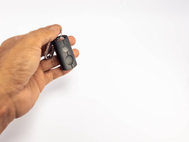 Yakın çekim, anahtarlığa bağlı küçük, siyah bir uzaktan kumandayı tutan bir el içerir. Kumandanın iki tuşu var, birinde güç sembolü var. Arka plan bembeyaz, temiz ve minimalist bir estetik sağlıyor. Görüntü iyi aydınlatılmış, yüksek