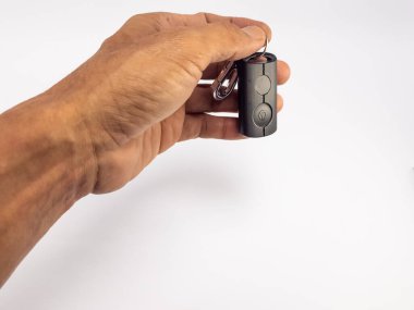 Yakın plan çekimde düz beyaz arka plana karşı parlak, koyu renkli bir uzaktan kumanda tutan bir el görülüyor. Kumandanın iki tuşu ve bir anahtarlığı var. Kompozisyon objeyi ve eli vurguluyor. Işıklandırma dokuları ve biçimi vurgular.