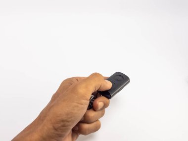 Bir kişinin elinde siyah bir uzaktan kumandayı tuşlarla tutarken, düğmeye basarken çekilmiş yakın plan bir fotoğraf. Resim, geniş bir kopyalama alanı olan temiz beyaz bir arkaplana göre ayarlanmış. Fotoğraf uzaktan kumandayı, el hareketini ve kontrol kavramını vurguluyor.