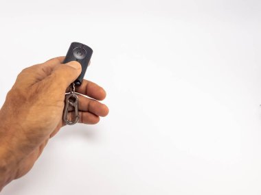 Siyah bir uzaktan kumandayı gümüş bir karabina tokasıyla tutan bir elin yakın çekimi. El, sade beyaz bir arka fonun tam ortasına yerleştirilmiştir. Görüntü iyi aydınlatılmış, elin dokusunu ve uzaktan kumandanın detaylarını vurguluyor