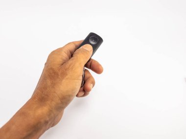 Yakından çekilen bir stüdyo resmi, siyah bir uzaktan kumandayı beyaz bir arka plana doğru tutan birinin elini yakalıyor. Görünür ten rengi ve dokusu olan el cihazın üzerindeki bir düğmeye basıyor. Görüntü, teknolojiyle etkileşimi vurguluyor.