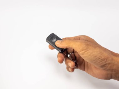 Yakın çekim, siyah bir uzaktan kumandayı sıkıca tutan bir eli gösteriyor. El, objeyi vurguluyor ve temiz, beyaz bir arkaplana dayanıyor. Uzaktan kumandanın ve elin dokusunun detaylarını vurguluyor. Kompozisyon 