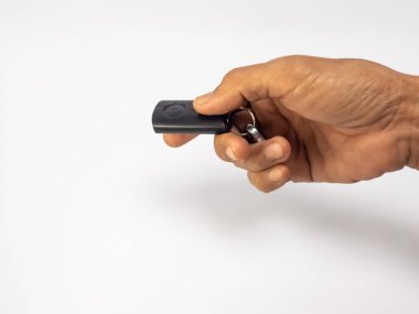 Stüdyoda, anahtarlığı olan siyah bir arabanın anahtarlığını tutan bir elin yakın plan fotoğrafı. El, düz beyaz bir arkaplanda konumlandırılmış. Görüntü, anahtarlığın parlak tasarımını ve otomotiv veya teknoloji için uygun kullanım kolaylığını vurgular
