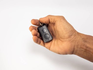 Yakından çekilen bir resimde siyah bir arabanın kumandasını tutan bir el görülüyor. Görünür deri dokusu ve damarları olan el, anahtarı nazikçe beşikte tutuyor. Arka plan temiz, beyaz bir yüzey. Uzaktan kumandayı ve el hareketini vurguluyor. Resim bir s iletir