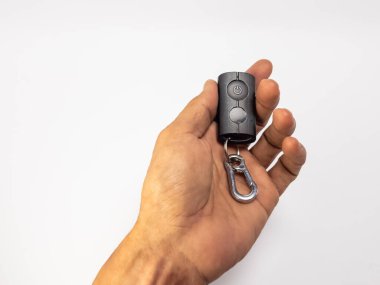 Siyah uzaktan kumandayla gümüş anahtarlık tutan birinin yakın plan fotoğrafı. Uzaktan kumandanın iki tuşu ve bir güç sembolü var. El, sade beyaz bir arka plana karşı, temiz ve minimalist bir estetik sağlıyor. Şey... 