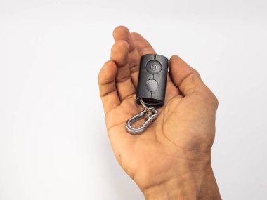 Bir insanın eli siyah bir uzaktan kumanda cihazını iki düğmesi ve gümüş bir karabina tokası ile beşikte tutuyor. Cihaz düz beyaz bir arkaplanda tutuluyor. Görüntü iyi aydınlatılmış, el dokusunu ve uzaktan kumandanın detaylarını gösteriyor. Basit.