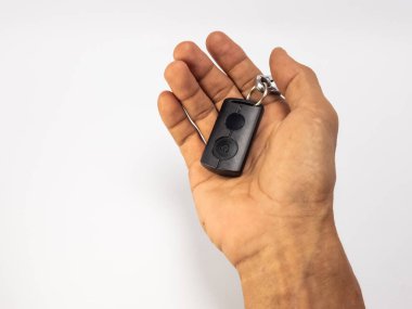 Yakın plan çekimde siyah bir arabanın anahtarlığını beyaz arka planda tutan bir el görülüyor. El kahverengi derili, görünürde detaylar var, doku ve damarlar gibi. Anahtarlık odak noktasında, düğmeleri ve yüzükleri gösteriyor. Kompozisyon basit, vurgula