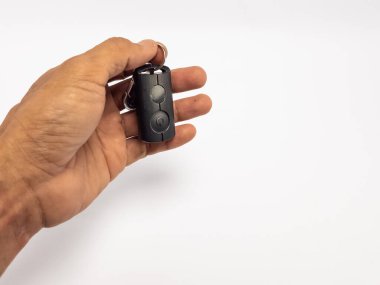 Yakın plan çekimde siyah bir arabanın anahtarlığını beyaz arka planda tutan bir insan eli görülüyor. Anahtarlıkta iki düğme ve bir anahtarlık var. El biraz bronzlaşmış ve anahtar düğmesine odaklanmış. Kompozisyon basit, vurgulanıyor.