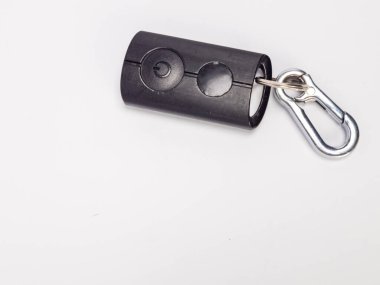 Yakın çekim, gümüş bir karabina şarjörüne bağlı iki düğmeli siyah bir uzaktan kumanda ve anahtarlık gösteriyor. Kumanda beyaz bir yüzeye yerleştirilmiş. Kompozisyon nesneye ve detaylarına odaklanır, temiz ve basit bir estetikle.