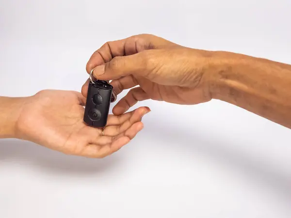 Yakın plan çekimde iki el arabanın anahtarlarını düz beyaz arka plan ile değiş tokuş ediyor. Bir el anahtarları sunarken, diğer el anahtarları alıyor. Bu görüntü sahiplik, transfer, satın alma ve otomotiv endüstrisi kavramlarını temsil ediyor. T